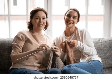 Head shot portrait smiling mature mother and grownup daughter knitting, sitting on cozy couch at home, happy young woman with middle aged mum looking at camera, enjoying leisure time together