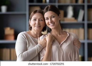 Head Shot Portrait Smiling Middle Aged Mother With Adult Grown Up Daughter Looking At Camera, Happy Young Woman With Older Mum Hugging, Standing In Living Room At Home And Posing For Family Photo