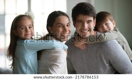 Head shot portrait smiling mother and father piggy backing adorable two kids, posing for funny family portrait at home, happy parents with little daughter and son looking at camera, having fun