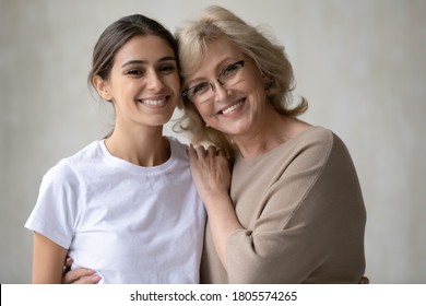 Head shot portrait of smiling multiracial different female generations family. Happy 60s european mother in eyeglasses cuddling shoulders of attractive young 30s arabic ethnicity grown up daughter.