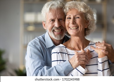 Head shot portrait smiling older wife and husband hugging, enjoying tender moment together, senior couple posing for family photo at home, caring loving mature man embracing woman from back