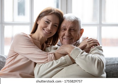 Head Shot Portrait Smiling Older Father And Grownup Daughter Looking At Camera, Sitting On Cozy Couch At Home, Overjoyed Young Woman And Mature Man Hugging, Cuddling, Two Generations Concept