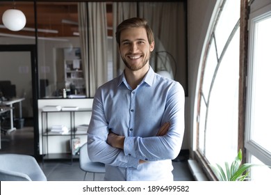 Head shot portrait smiling successful young businessman standing with arms crossed in modern office, happy executive boss startup founder looking at camera, posing for corporate photo alone