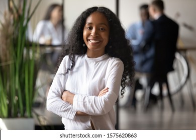 Head shot portrait of smiling young african american female professional, standing with folded hands at modern office. Happy mixed race businesswoman entrepreneur team leader posing for photo.