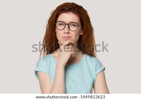 Head shot portrait of thoughtful millennial red-haired woman in eyewear looking suspiciously at camera, touching chin, doubtful, making important decision, isolated on white grey studio background.