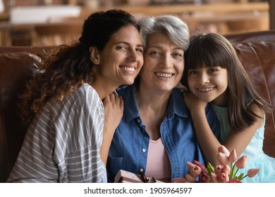 Head Shot Portrait Three Generations Of Women Celebrating Mature Grandmother Birthday Or 8 March At Home, Grownup Daughter And Little Granddaughter Presenting Gift And Flowers To Older Woman