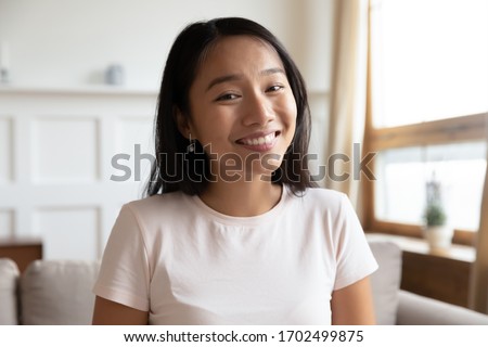Head shot portrait Vietnamese millennial attractive woman sit on couch in living room alone, smiling looking at camera webcam, make video conference call distant interaction using modern tech concept
