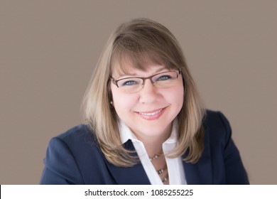 Head Shot Of Professional Business Woman In Her Upper Forties With Blonde Hair And Blue Eyes Wearing A Blue Suit Jacket, White Blouse, Necklace, Earrings And Glasses. Horizontal Landscape Orientation.