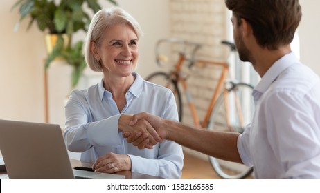 Head Shot Satisfied Middle Aged Businesswoman Shaking Hands With Young Male Partner. Smiling Older Female Professional Making Agreement With Client At Meeting. Happy Boss Praising Colleague.