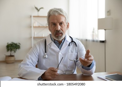 Head shot serious mature doctor wearing white uniform coat with stethoscope speaking, looking at camera, gesturing, senior therapist practitioner gp consulting patient online, making video call