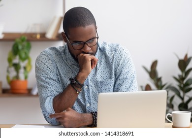 Head shot serious puzzled African American businessman looking at laptop screen sitting in office. Executive managing thinking received bad news keeping fist at chin waiting hoping positive result