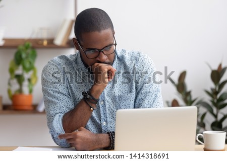 Head shot serious puzzled African American businessman looking at laptop screen sitting in office. Executive managing thinking received bad news keeping fist at chin waiting hoping positive result