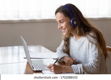 Head shot side view smiling young mixed race girl wearing wireless headphones, looking at laptop screen, watching educational video online. Happy millennial woman enjoying studying remotely at home.
