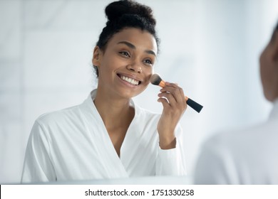 Head shot smiling African American woman applying powder or blush on cheeks, using cosmetics brush, looking in mirror, beautiful girl wearing bathrobe putting daily makeup, standing in bathroom