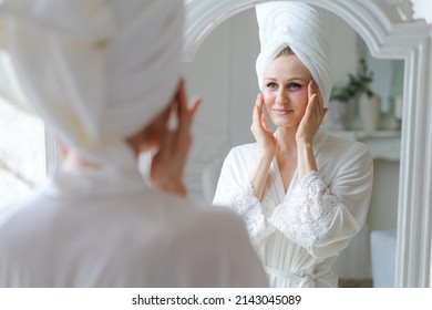 Head shot Smiling attractive female caucasian woman applying hydrogel head patches further under eye area looking at mirror enjoying daily routine against margin in robe and towel after morning shower