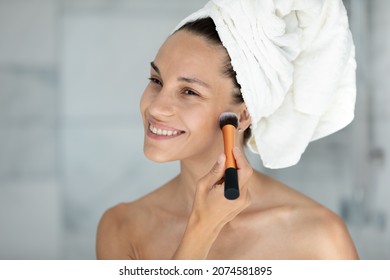 Head shot smiling attractive young mixed race hispanic woman applying foundation with brush after morning shower, getting ready doing nude flawless make up in bathroom, beauty routine concept.
