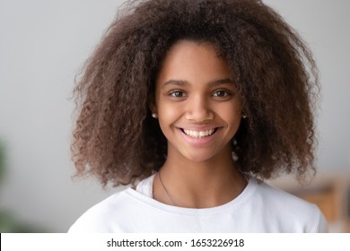 Head shot of smiling cute teenage african american girl with afro hair looking at camera at home, happy beautiful black teenager posing indoor, pretty adolescent mixed race teen school child portrait