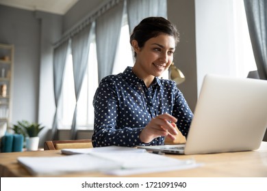 Head shot smiling pretty indian girl sitting at table, looking at laptop screen. Happy hindu ethnic woman reading message email with good news, chatting with clients online, working remotely.