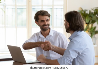 Head shot smiling young businessman shaking hands with confident male coworker, thanking for help with computer software explanation or praising for good team work, sitting together at office. - Shutterstock ID 1582165570