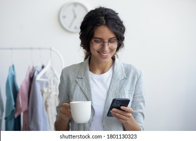 Head Shot Smiling Young Businesswoman Holding Cup Of Tea Coffee, Consulting Clients Online Using Mobile Application. Happy Millennial Creative Designer Stylist Enjoying Pause Break Time In Office.