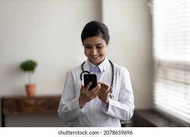 Head Shot Smiling Young Indian Female Doctor Physician In White Uniform With Stethoscope Using Smartphone, Standing In Medical Office, Nurse Or Therapist Typing And Looking At Phone Screen