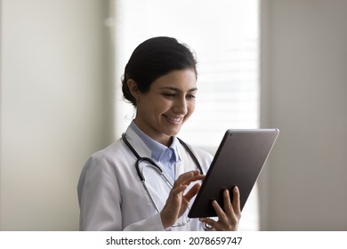 Head Shot Smiling Young Indian Woman Doctor In White Uniform With Stethoscope Using Tablet Close Up, Typing, Looking At Modern Electronic Device Screen, Physician Therapist Consulting Patient Online