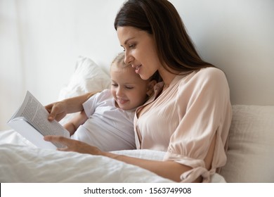 Head Shot Smiling Young Mommy Lying In Bed Under A Blanket With Little Cute Daughter, Reading Fairytales In Morning. Small Adorable Preschool Kid Girl Looking In Book, Listening To Stories In Bedroom.