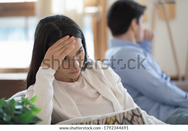Head shot stressed confused young mixed race\
woman sitting in sofa separately with offended husband, ignoring\
each other after conflict quarrel, marriage relationship\
misunderstanding problem.