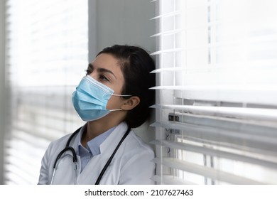 Head shot stressed unhappy young Indian doctor or nurse in protective facemask medical respirator leaning head against wall sill in clinic office room, feeling exhausted alone after hard workday.