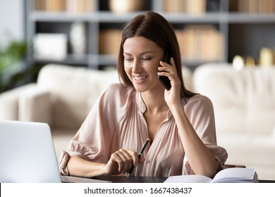 Head shot successful businesswoman working remotely at home, looking at computer screen, talking on smartphone with client. Smiling young woman chatting with friend on mobile phone during break time.