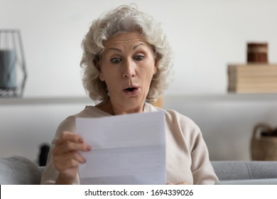 Head Shot Surprised Middle Aged Mature Woman Looking At Paper Letter, Reading Unbelievable News Notification, Sitting Alone On Sofa At Home. Stressed Older Grandmother Received Bank Account Balance.