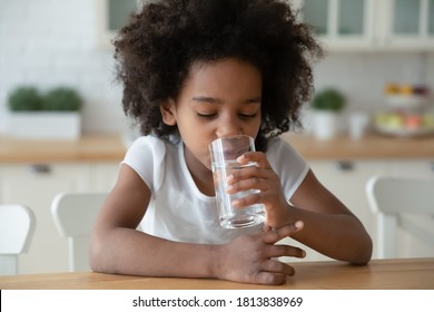 Head Shot Thirsty Small Curly African American Little Child Girl Drinking Glass Water, Hydrating Organism Indoors. Happy Little Mixed Race Kid Enjoying Morning Healthcare Routine Alone In Kitchen.