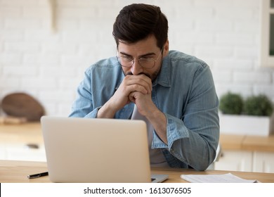 Head shot thoughtful businessman in eyewear looking at computer screen, sitting at table at home. Pensive confused young man thinking of problem solution stuck with task, working with laptop remotely. - Shutterstock ID 1613076505