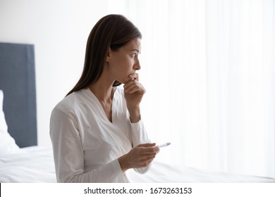 Head Shot Thoughtful Young Woman In Nightgown Bathrobe Sitting On Bed, Holding Quick Plastic Test In Hands, Worrying About Infertility Or Unwanted Unplanned Pregnancy, Future Maternity Concept.