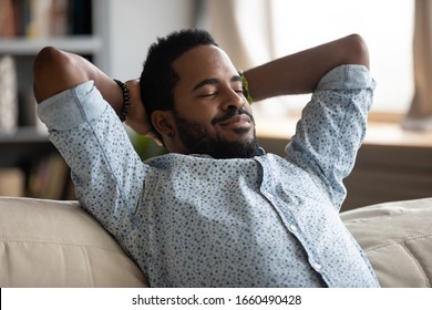 Head Shot Tranquil Happy Young African Ethnicity Man Folded Hands Behind Head, Relaxing On Comfy Sofa, Daydreaming Sleeping At Home. Peaceful Carefree Biracial Guy Alone Napping On Couch Indoors.