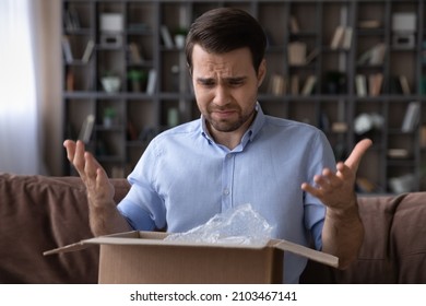 Head shot unhappy dissatisfied man opening parcel at home, sitting on couch with cardboard box, angry displeased customer confused by wrong or damaged order, bad delivery shipping service concept