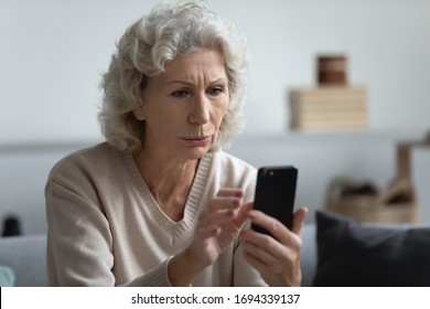 Head Shot Worried Senior Mature Retired Woman Holding Phone, Reading Unpleasant News In Social Media. Unhappy Older Middle Aged Grandmother Sitting On Sofa, Received Bad News Message On Smartphone.