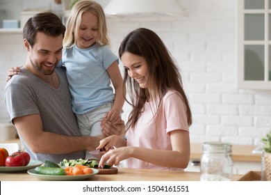 Head shot young handsome father holding little adorable happy daughter, watching smiling millennial mommy chopping fresh vegetables on wooden board, preparing healthy lunch dinner in kitchen at home.