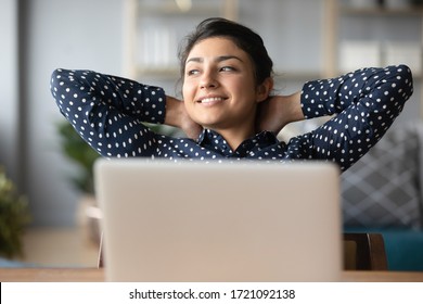 Head shot young indian woman crossed hands behind head, enjoying break time at home. Peaceful carefree millennial hindu student resting at table with computer, looking aside, dreaming of future.