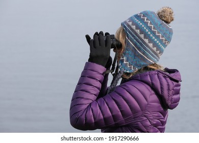 A Head And Shoulder Image Of A Lady Looking Through Binoculars.She Wears Gloves A Bobble Hat And A Down Jacket.Plain Background Of Sea.