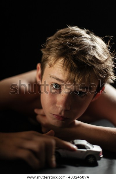 Head and Shoulders Close
Up of Young Shirtless Teenage Boy with Head Resting on Hands While
Lying on Floor with Toy Car in Studio with Black Background and
Copy Space