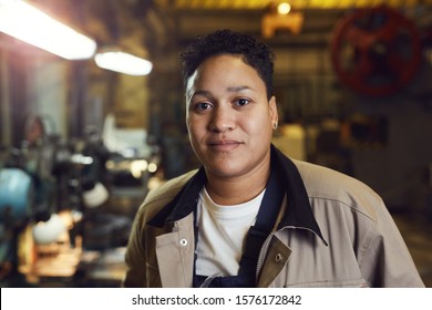 Head and shoulders portrait of contemporary mixed-race woman posing in factory workshop at looking at camera, copy space