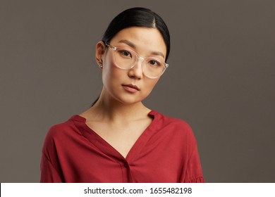 Head And Shoulders Portrait Of Elegant Asian Woman Wearing Glasses And Looking At Camera While Standing Against Grey Background In Studio, Copy Space