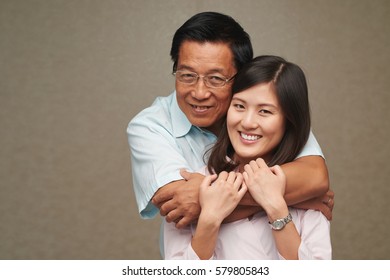 Head And Shoulders Portrait Of Mature Asian Man Standing Behind His Pretty Daughter And Hugging Her Tenderly While They Posing For Photography