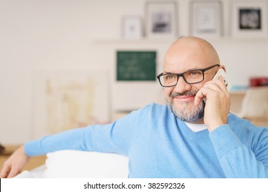 Head and Shoulders Portrait of Mature Man with Gray Facial Hair Wearing Eyeglasses and Casual Blue Shirt Smiling While on Cell Phone in Living Room on Relaxing Day at Home, Copy Space to Left of Image
