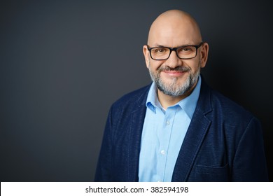 Head And Shoulders Portrait Of Mature Man With Facial Hair Wearing Eyeglasses And Dress Shirt With Denim Jacket Smiling Warmly At Camera In Studio With Dark Gray Background And Copy Space