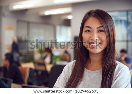 Head And Shoulders Portrait Of Smiling Young Asian Businesswoman  Working In Busy Modern Office