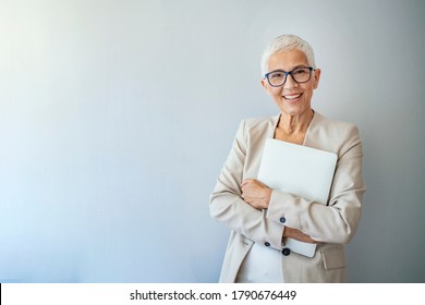 Head And Shoulders View Of Beautiful Older Woman With Short Grey Hair And Beige Jacket Against Grey Background. Business Woman With Glasses Smiling. Portrait Of Beautiful Mature Woman