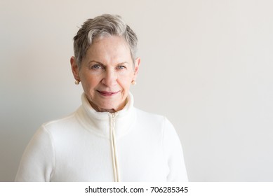 Head And Shoulders View Of Beautiful Older Woman With Short Grey Hair And Blue Eyes In Cream Jacket Against Neutral Wall (selective Focus)