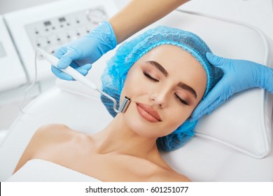 Head And Shoulders Of Woman Lying On Couch In Blue Cap In Cosmetological Clinic With Closed Eyes. Doctor's Hands In Blue Gloves Holding Electrode For Galvanization Near Her Face